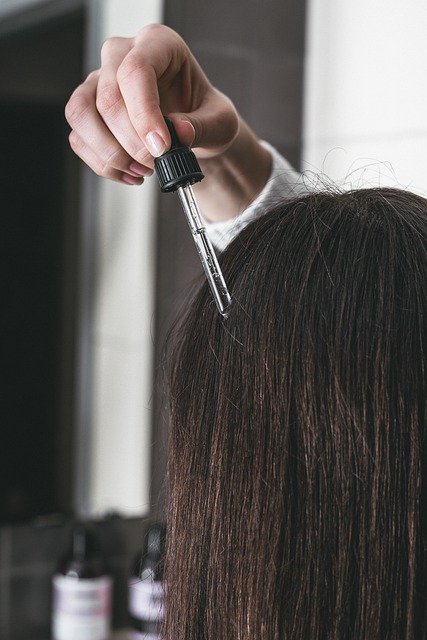 Person applying hair serum or oil to scalp using a dropper on straight, dark hair