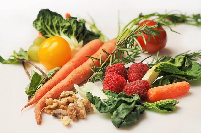 A colorful assortment of fresh vegetables and fruits including carrots, raspberries, spinach, tomato, yellow cherry tomato, and walnuts on a white background.