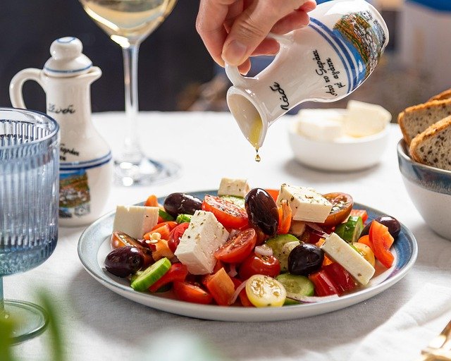 A close-up of a fresh Greek salad with tomatoes, cucumbers, olives, and feta cheese, being drizzled with olive oil from a small decorative jug.
